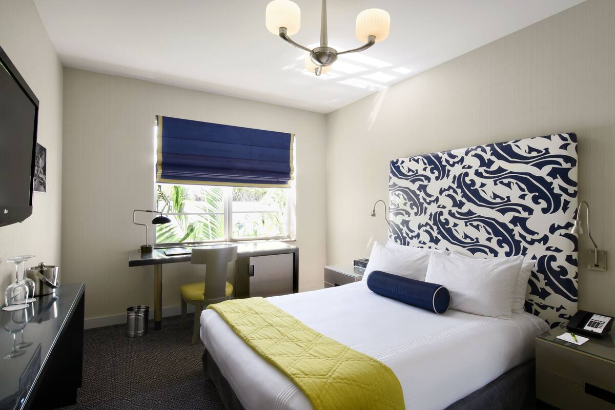 A hotel room with a bed, desk, chair, and a window with a blue blind.