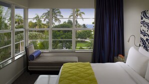 Una camera d'albergo con un letto, una panca, un'ampia finestra vista palme e l'oceano, e un lampadario.