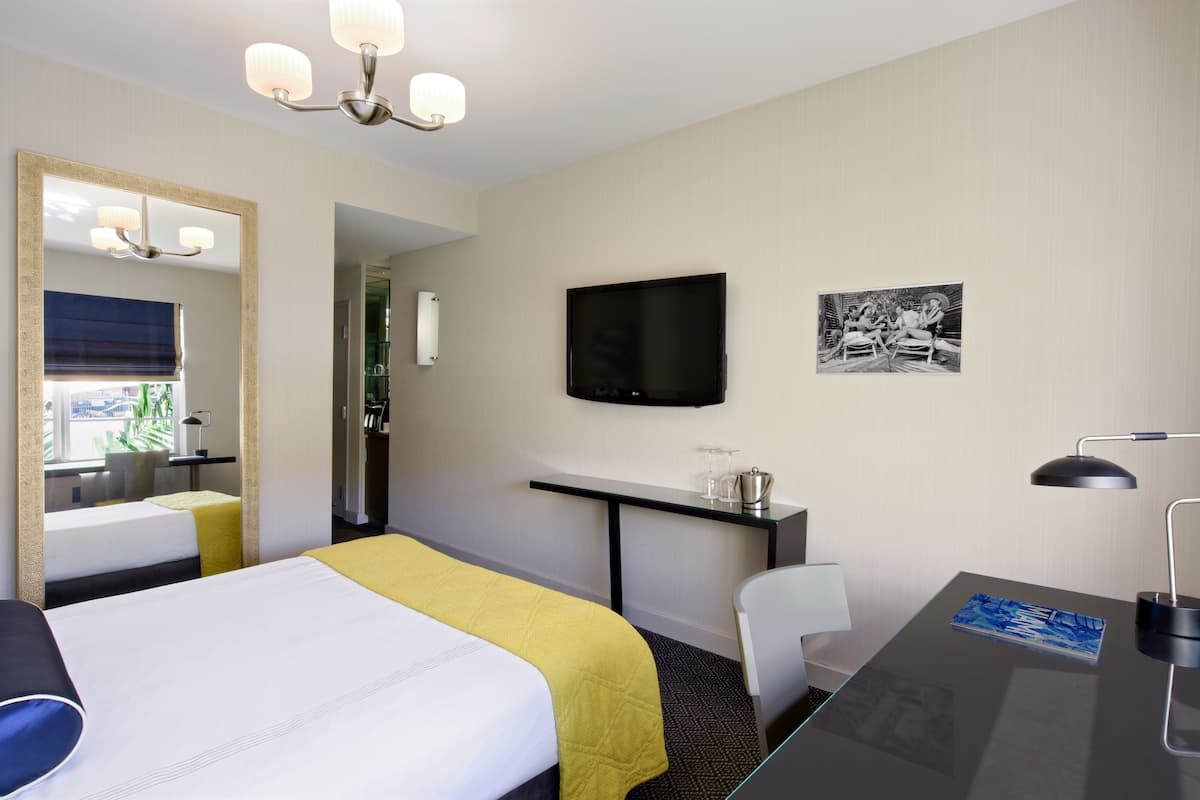 A hotel room with a bed, a desk, a chair, a television, a mirror, and artwork on the wall.
