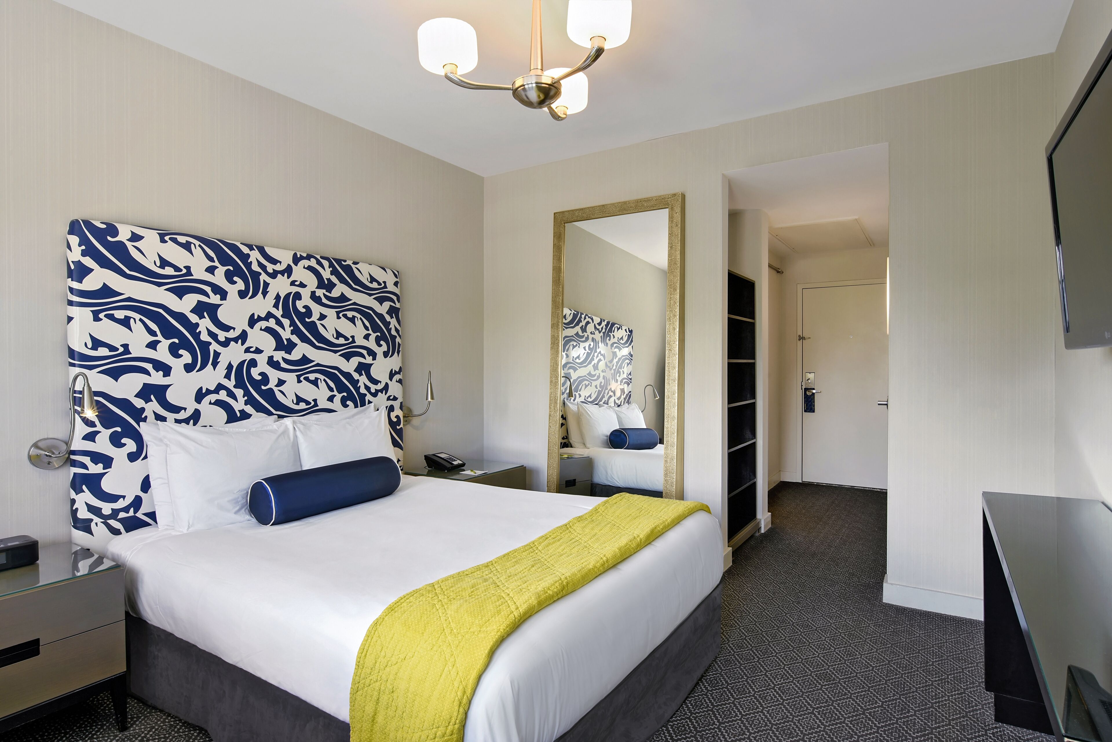 A hotel room with a large bed, a bedside table, a nightstand, a desk, a chair, a mirror, a wardrobe, and a television.