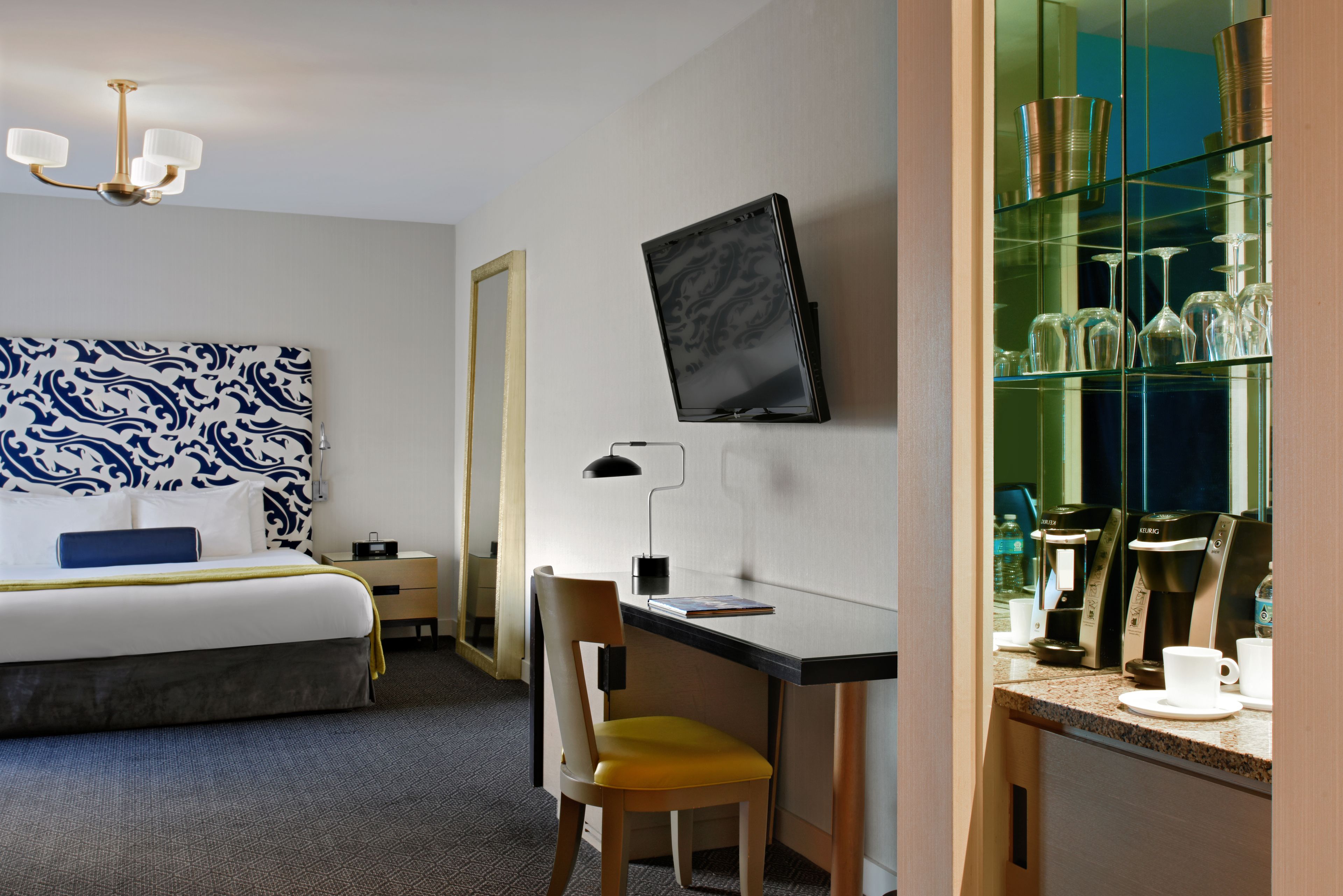 Habitación de hotel con cama, escritorio, silla, cafetera y vitrina de cristal.