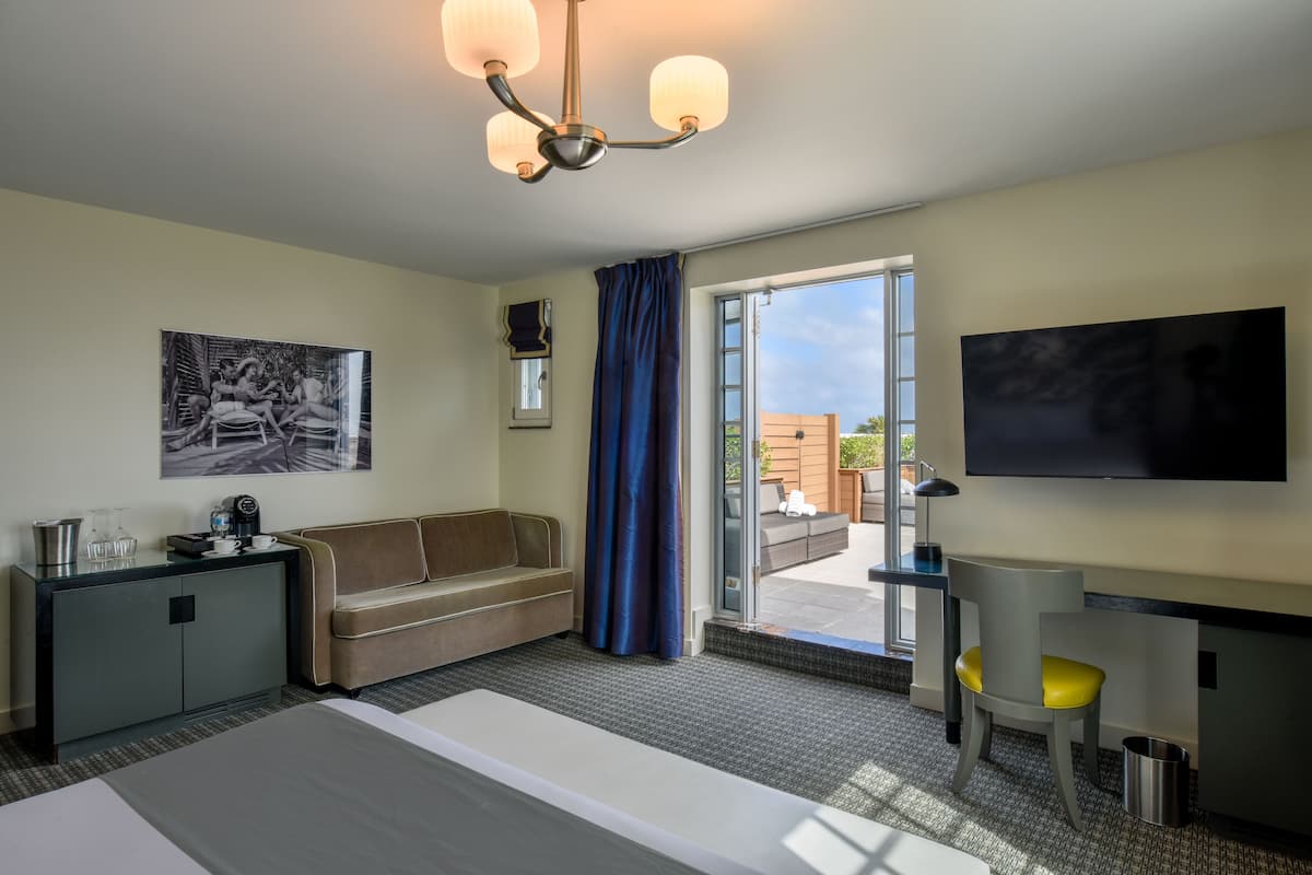 A hotel room with a bed, a desk, a chair, a sofa, a television, a painting, a window with curtains, and a balcony.