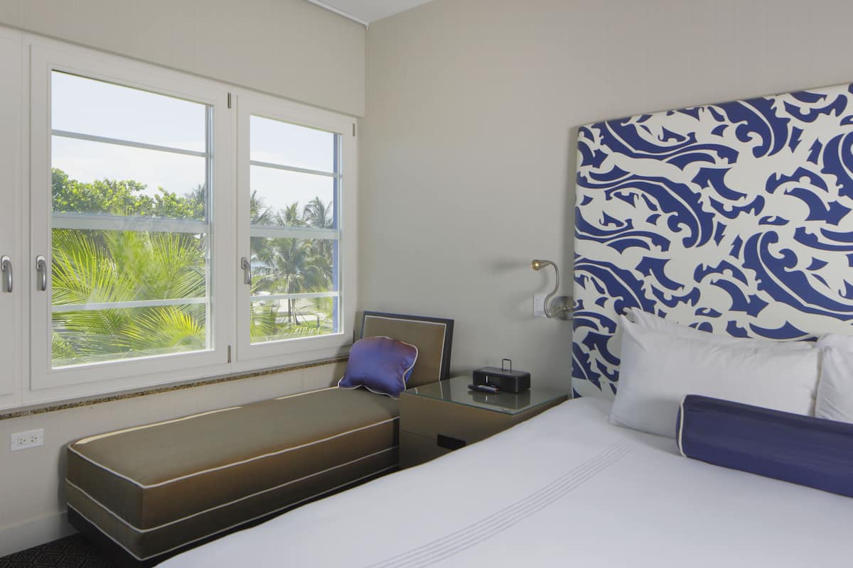 A hotel room with a bed, a bench, a bedside table, and a window with a view of greenery.