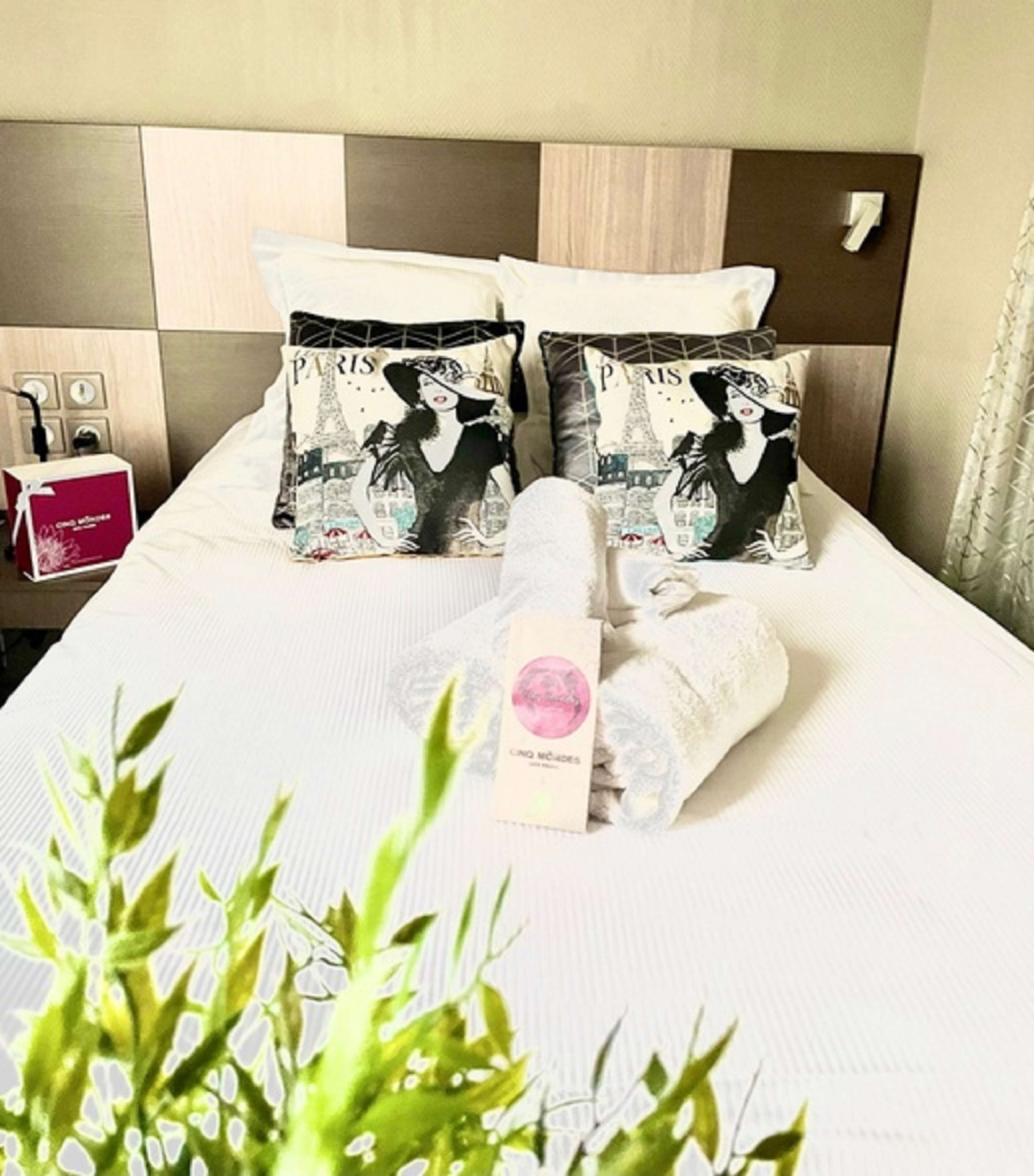 Un lit bien fait, agrémenté de deux coussins décoratifs aux motifs parisiens, une serviette roulée et un panneau de commande de la chambre bien visible.