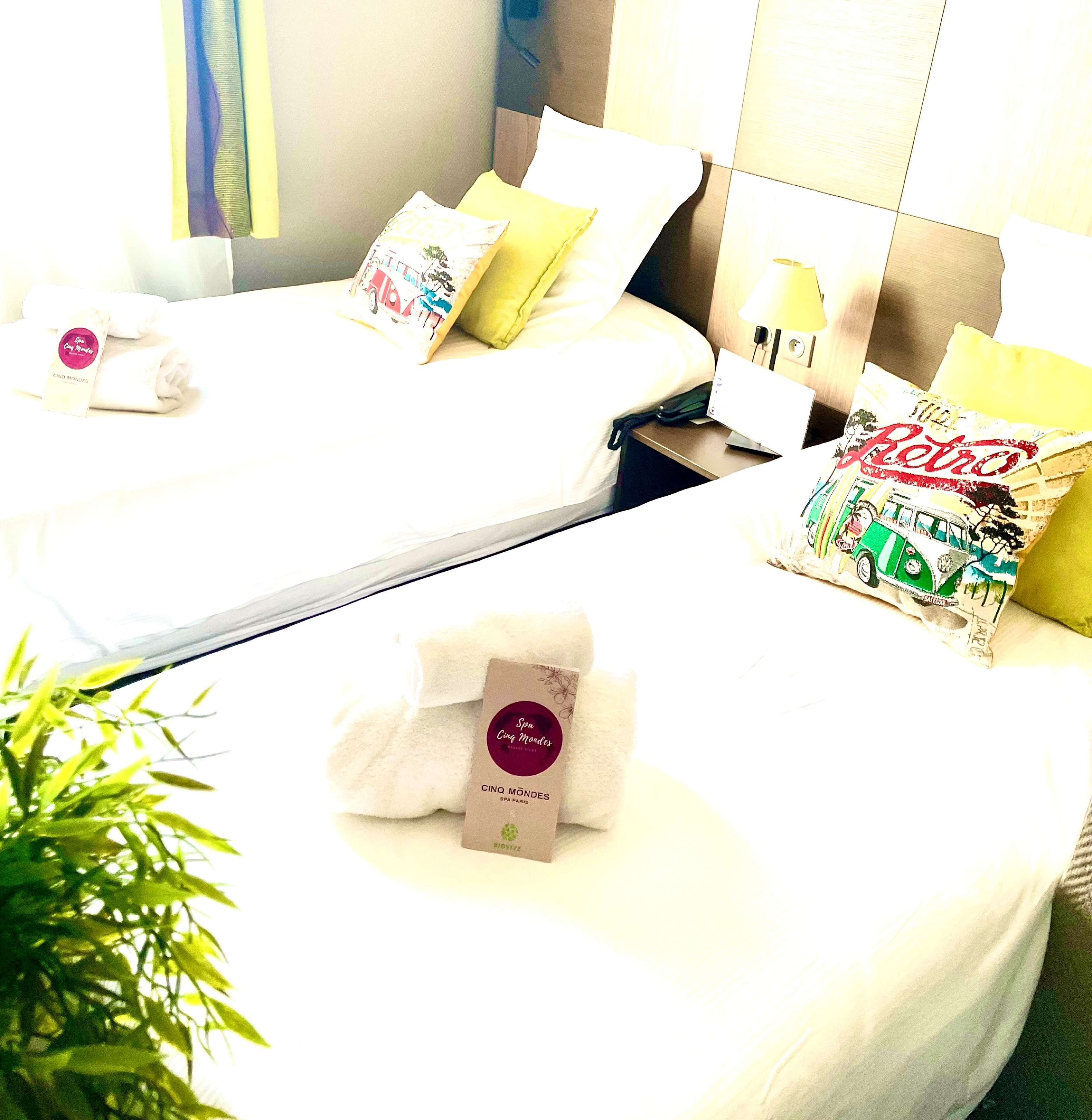 A hotel room with two beds, a bedside table with a lamp, and a plant.