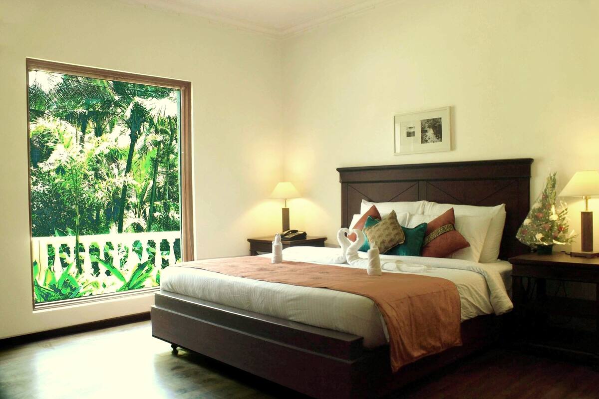 A bedroom with a large window, a bed with pillows and a blanket, two bedside tables with lamps, and a framed picture on the wall.