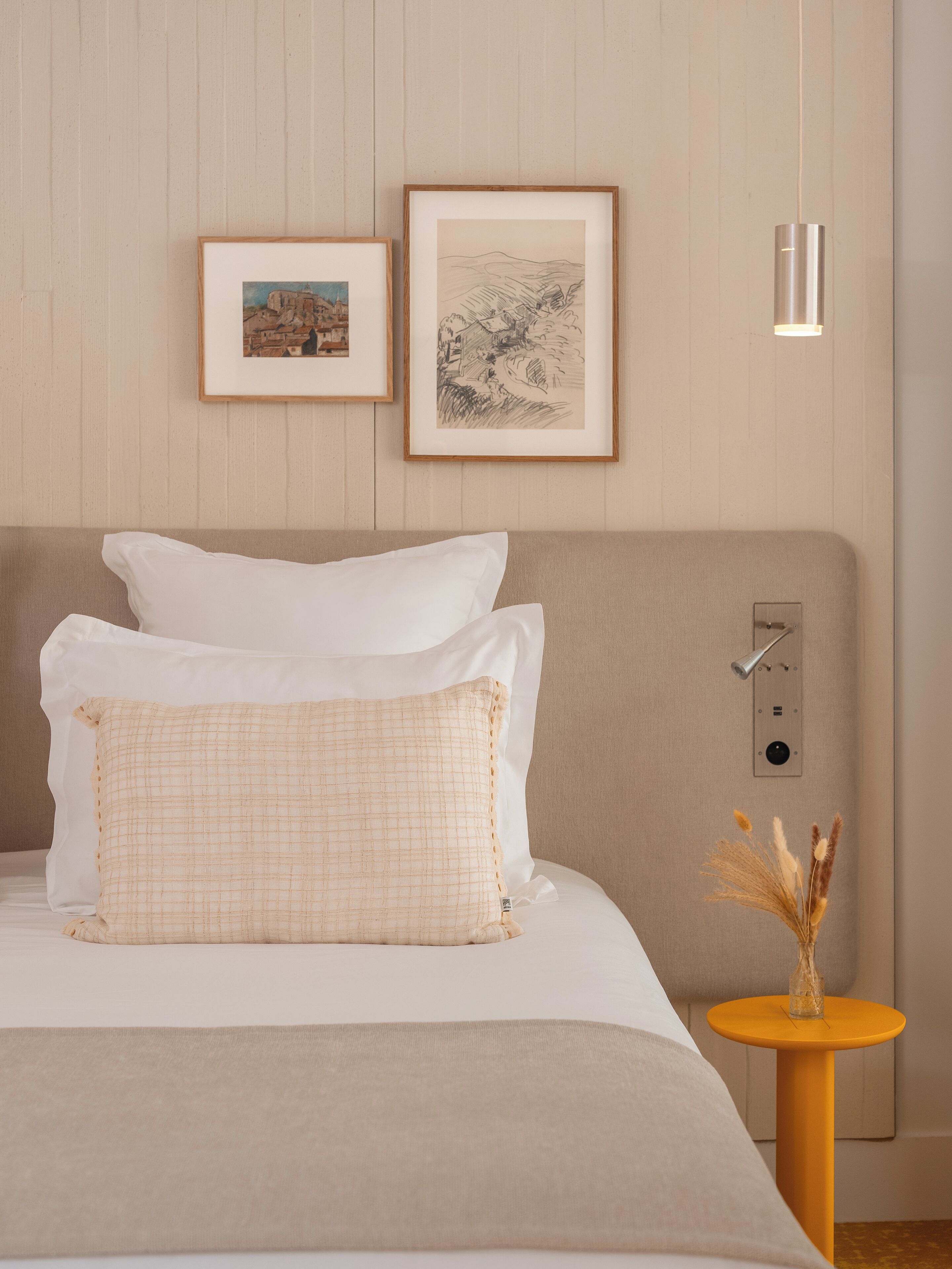 A neatly made bed with white and beige bedding, two framed artworks on the wall, and a bedside table with a vase of dried plants.