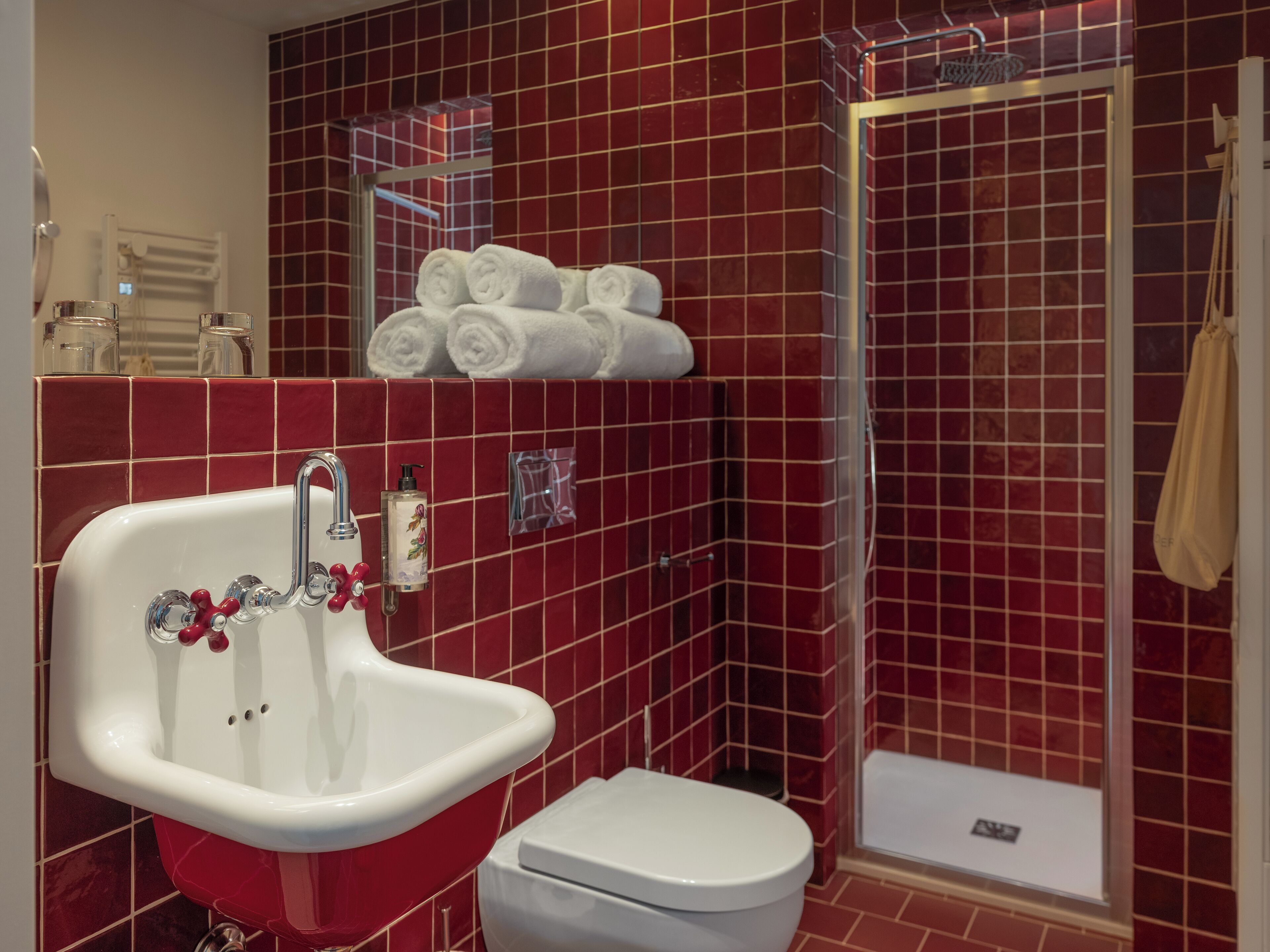 Casa de banho com parede revestida a azulejos vermelhos, lavatório branco com puxadores vermelhos, sanita branca e área de duche.