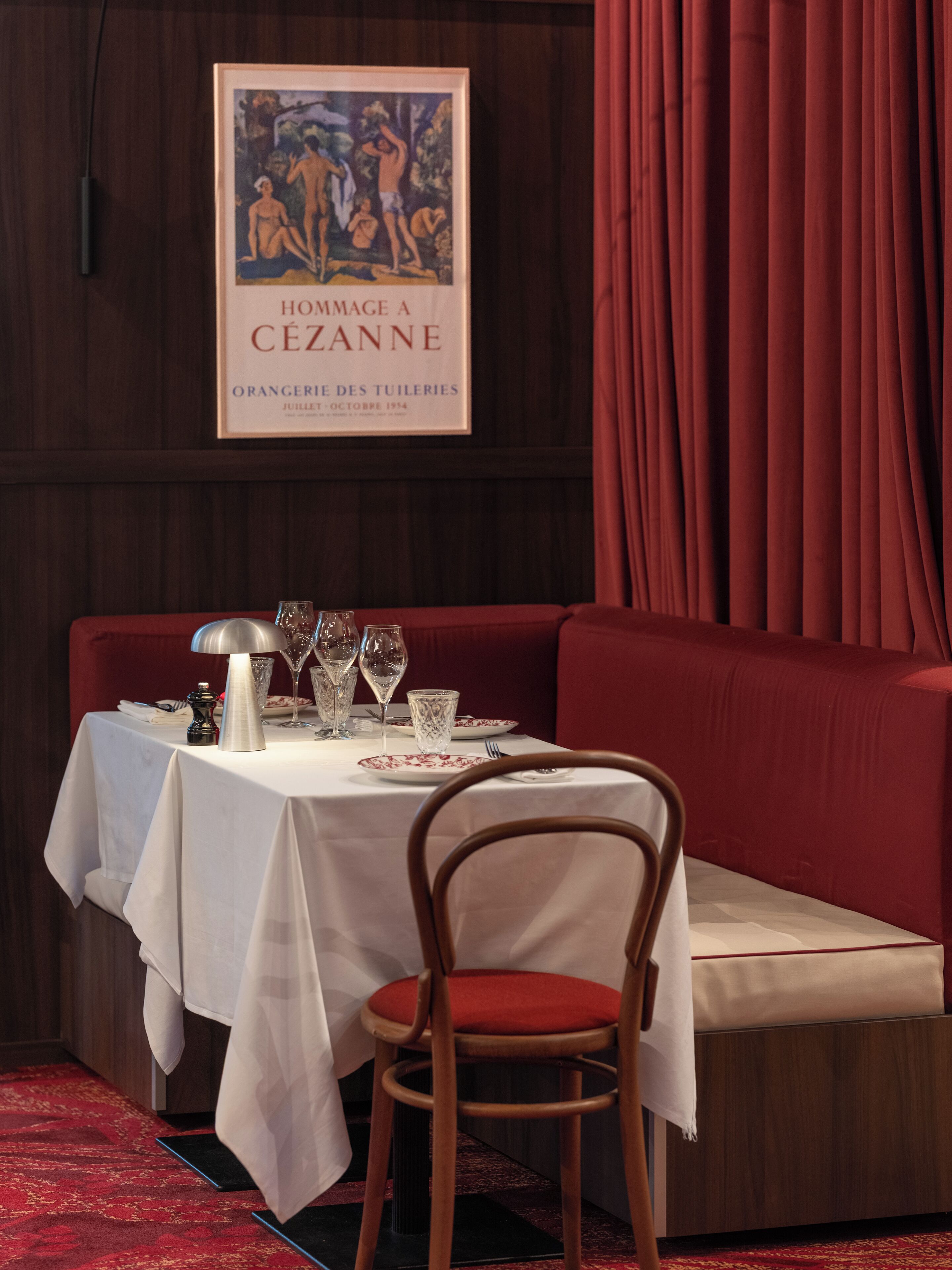 A dining area with a table set for two, a framed artwork above, and a red curtain to the right.
