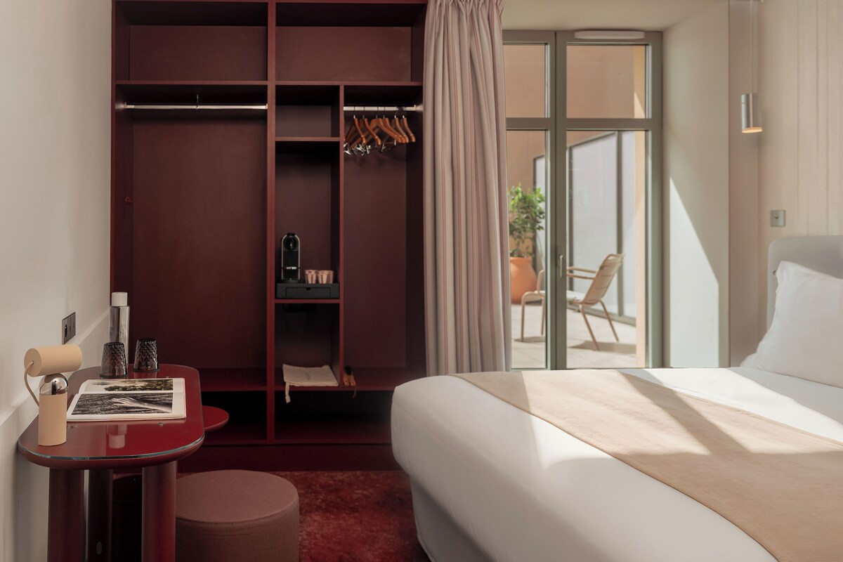 A hotel room with a bed, a desk with a chair, a wardrobe, and a sliding glass door to a balcony.