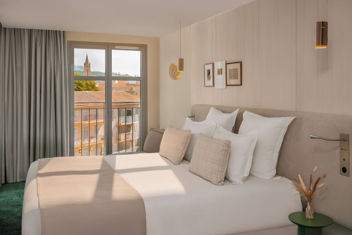 A hotel room with a large bed, a view of buildings through a window, and a green side table with a vase.