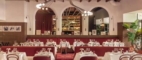 Un restaurante con varias mesas dispuestas para comer, vigas de madera en el techo y una zona de bar visible a través de un arco.