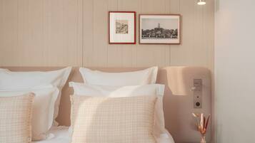 Un lit bien fait, avec du linge de lit blanc et des coussins à motifs, une petite table rouge surmontée d’un vase, et deux tableaux encadrés accrochés au mur.