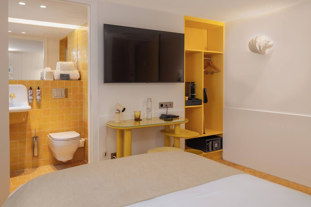 A modern bathroom with a toilet, sink, and mirror. There is a yellow vanity with a mirror, a small table, and a shelf with toiletries. A black TV is mounted on the wall above the vanity.