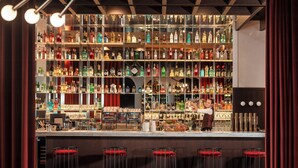 Zona de bar con un cantinero, estantes llenos de botellas de licor y taburetes rojos.