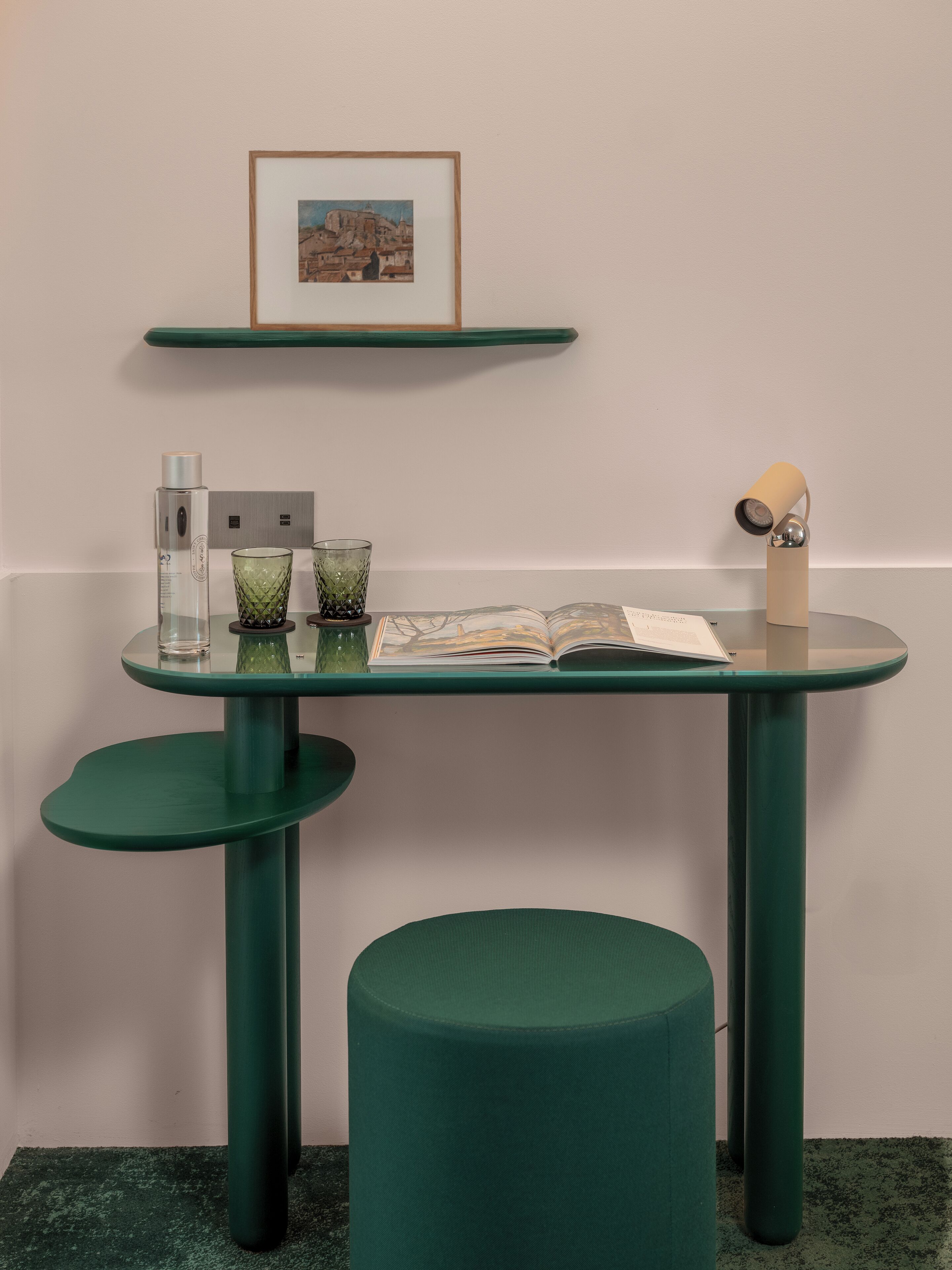 Una mesa color turquesa con tapa de vidrio, un cuadro enmarcado encima y un otoman verde al frente.