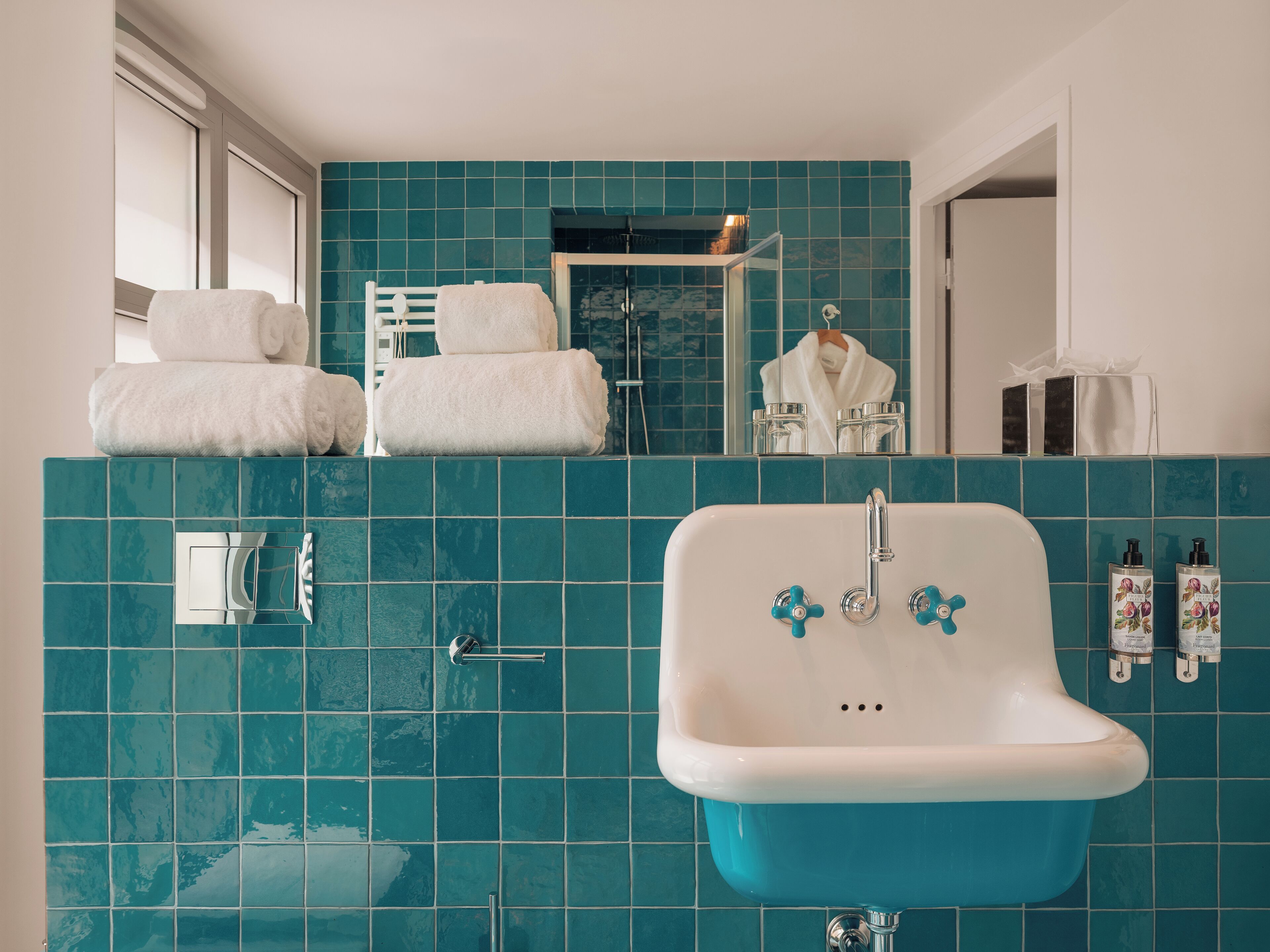 A bathroom with teal tiles, a white sink with chrome fixtures, and a mirror reflecting a towel rack and a shower area.