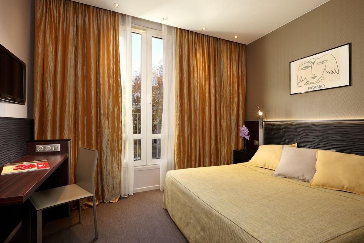 Habitación de hotel con cama, escritorio, silla, televisor y ventana con cortinas.