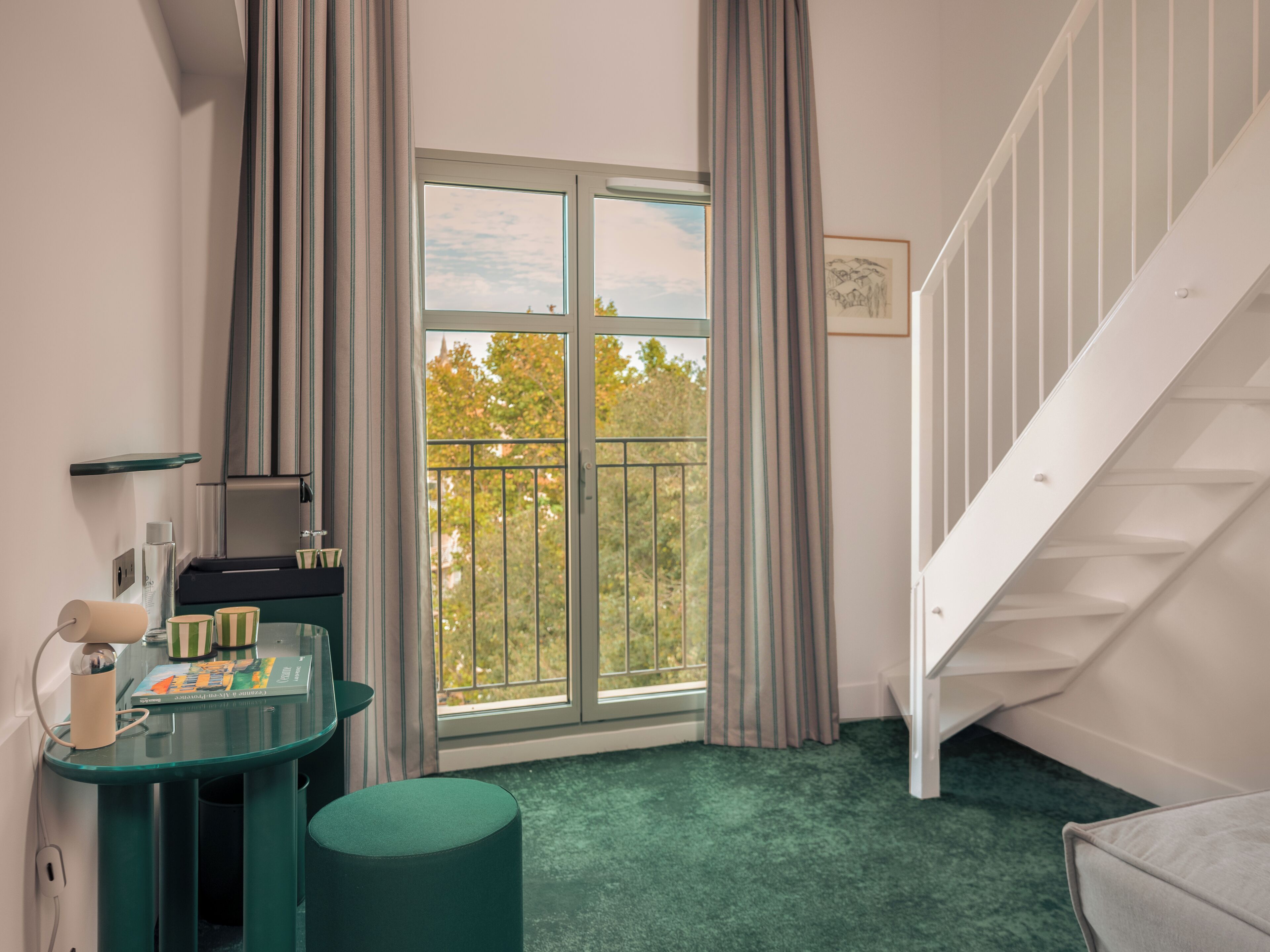 A room with a window, a desk, a chair, a green ottoman, and a white staircase.