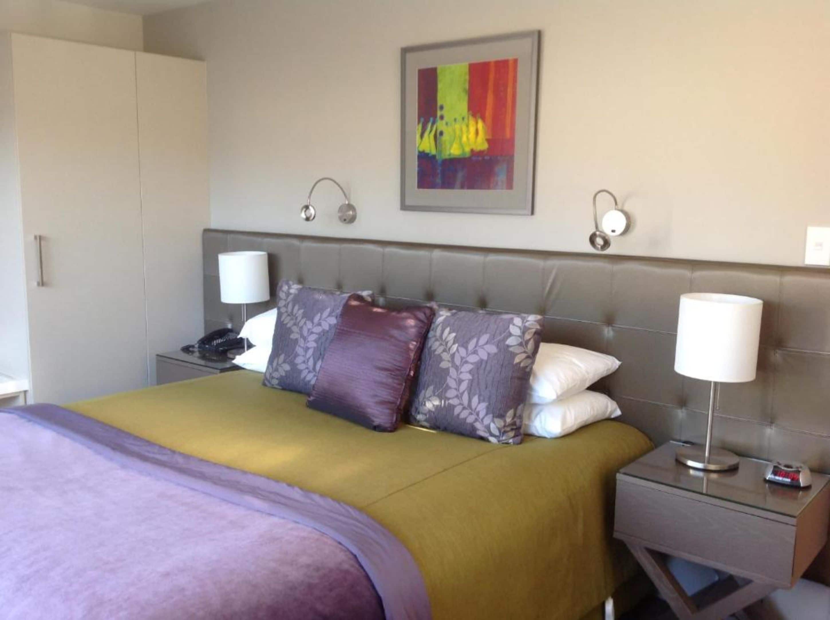 Una habitación de hotel con cama, mesita de noche, lámpara, teléfono y un cuadro en la pared.