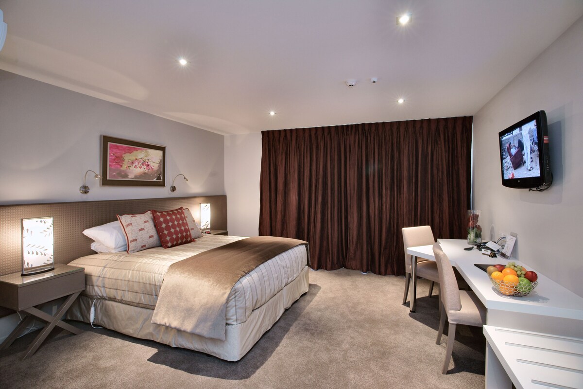 A hotel room with a bed, bedside table, desk, chair, and a TV on the wall.