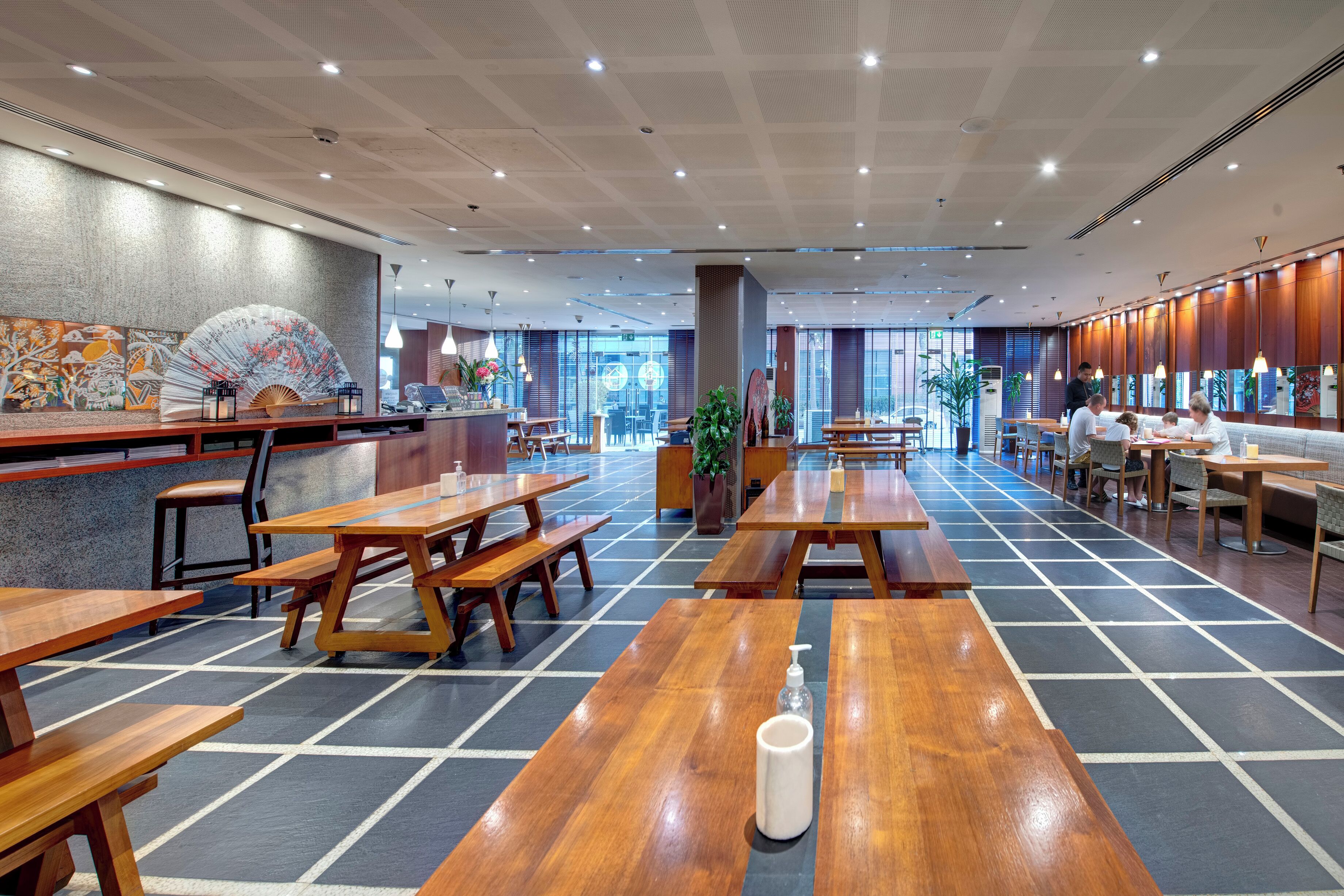 Un vaste espace de restauration intérieur, doté de tables et de bancs en bois, d’un grand ventilateur et offrant une vue sur l’extérieur grâce à de grandes fenêtres.
