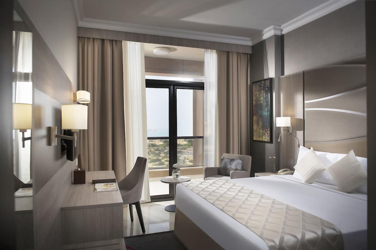 A hotel room with a large bed, a desk with a lamp, a chair, a small table with a vase, and a painting on the wall.