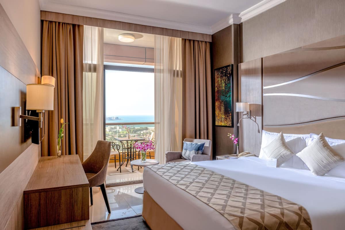 A hotel room with a large bed, a desk, a chair, a small table, and a balcony with a view.