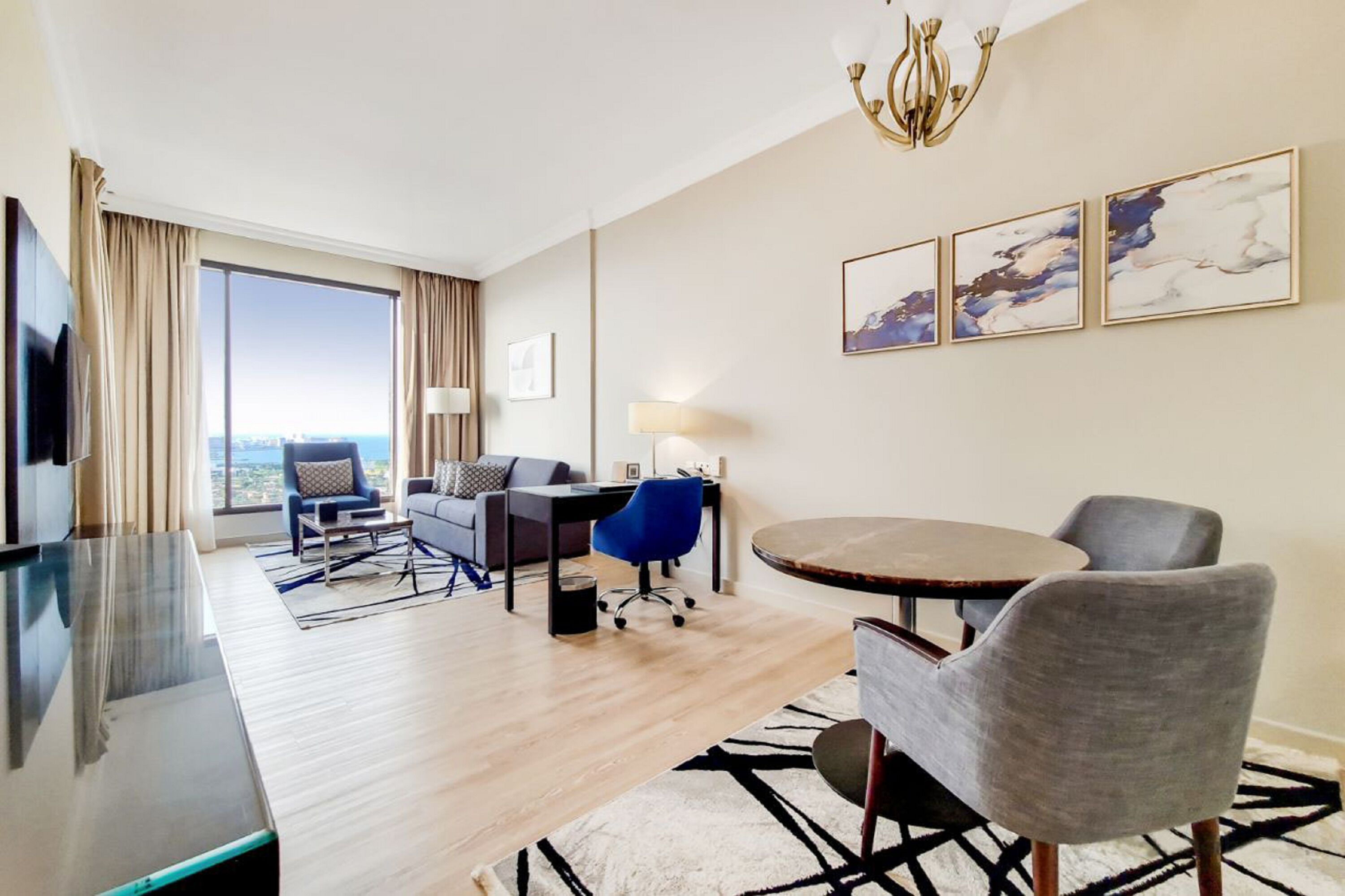Habitación de hotel con un ventanal grande, una mesa de comedor, un sofá, una silla azul y obras de arte en la pared.