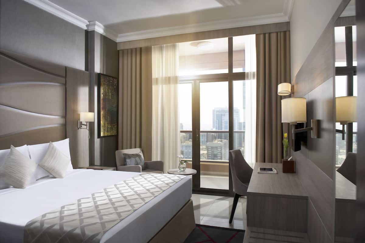 A modern hotel room with a large bed, a desk, a chair, and a view of the city through large windows.