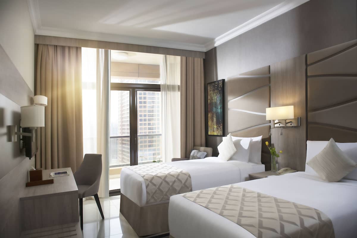 A hotel room with two beds, a desk, a chair, a lamp, and a view of the city.