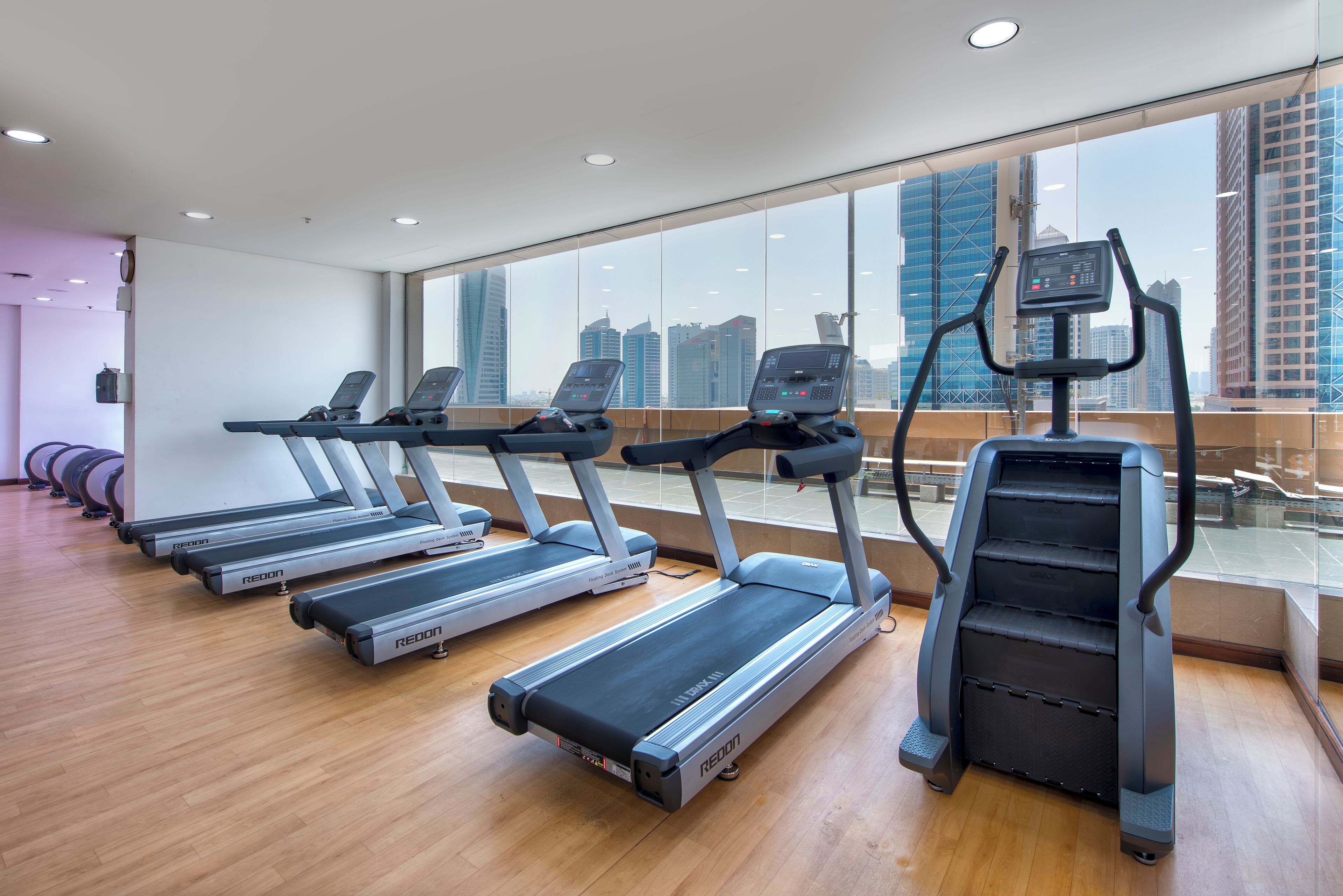 Ein Fitnessstudio mit Laufbändern und Crosstrainern, großen Fenstern mit Stadtblick und Holzboden.