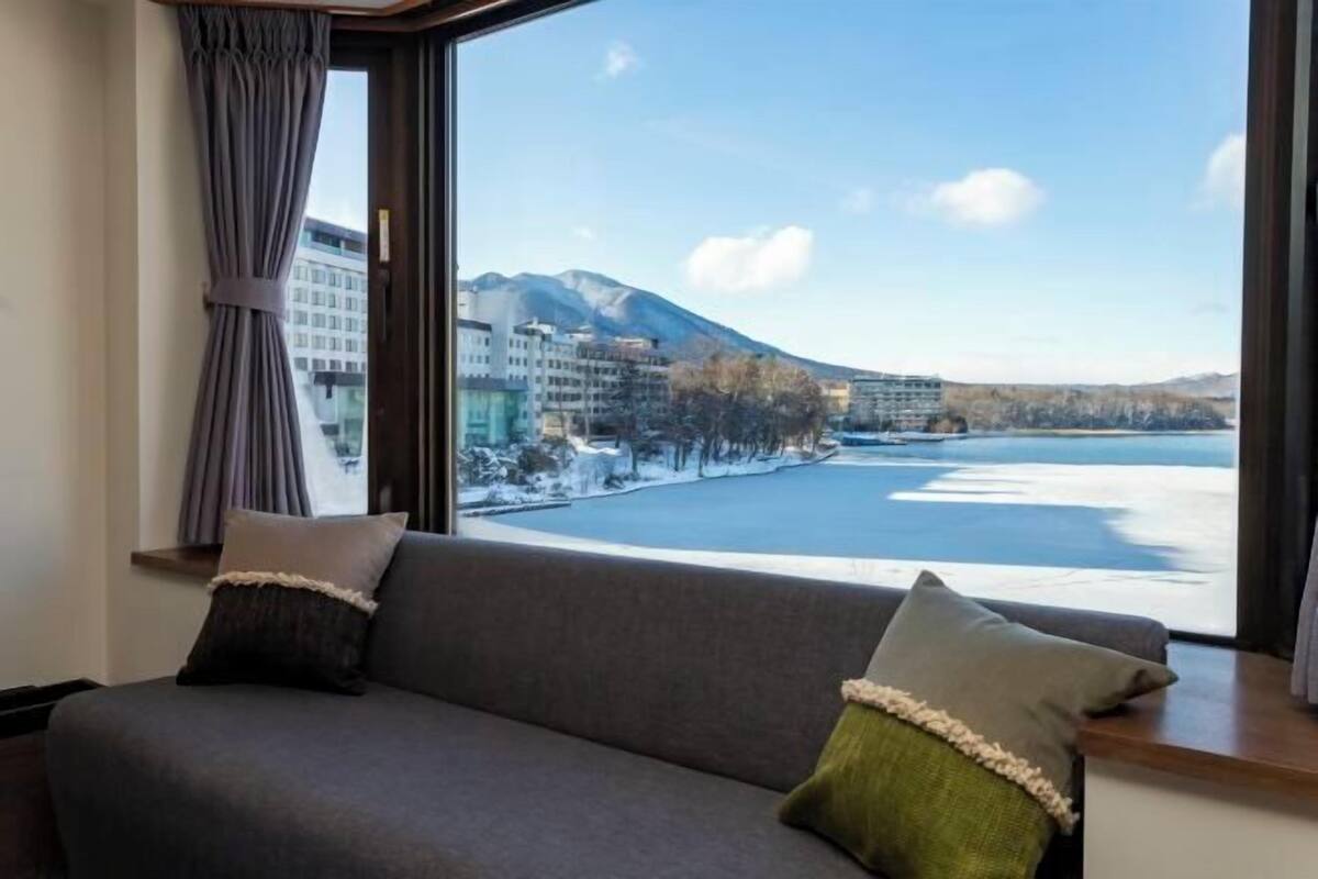 A room with a sofa, two pillows, a window with curtains, and a view of a snowy landscape with buildings and a mountain.