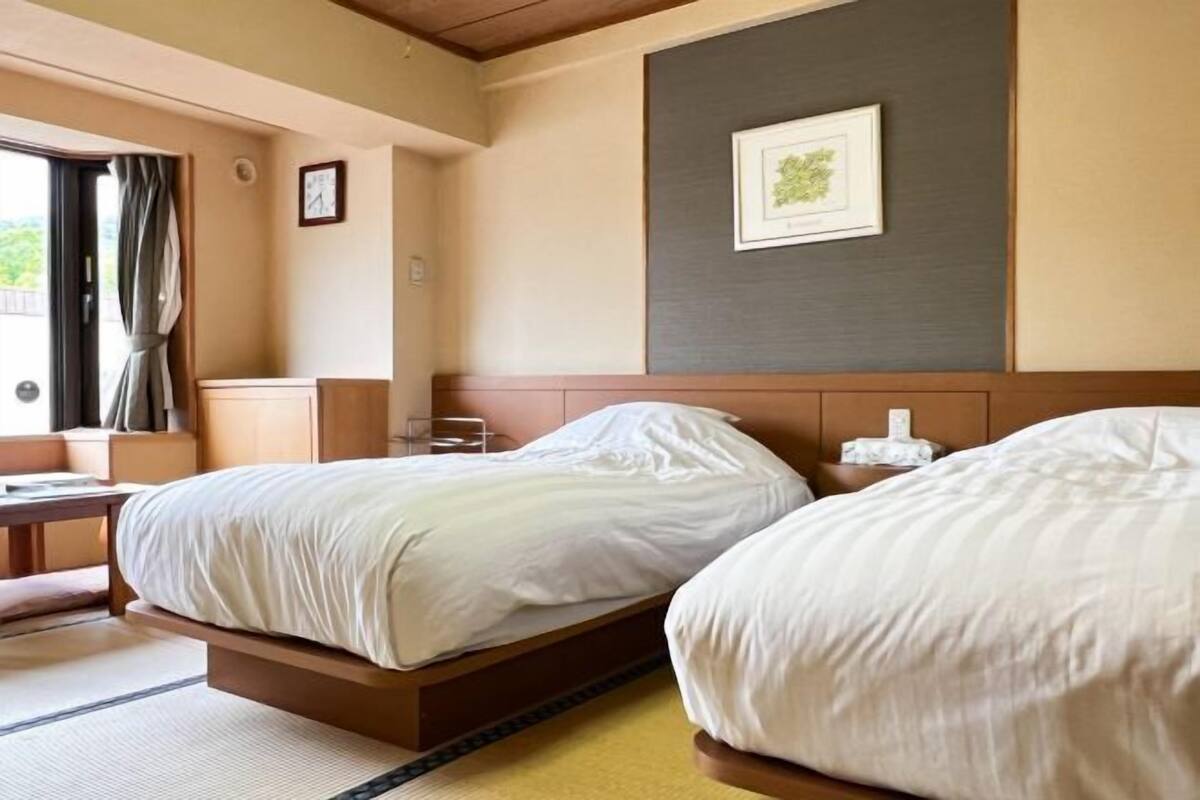 A hotel room with two beds, a wooden headboard, a framed picture on the wall, and a view of greenery outside.