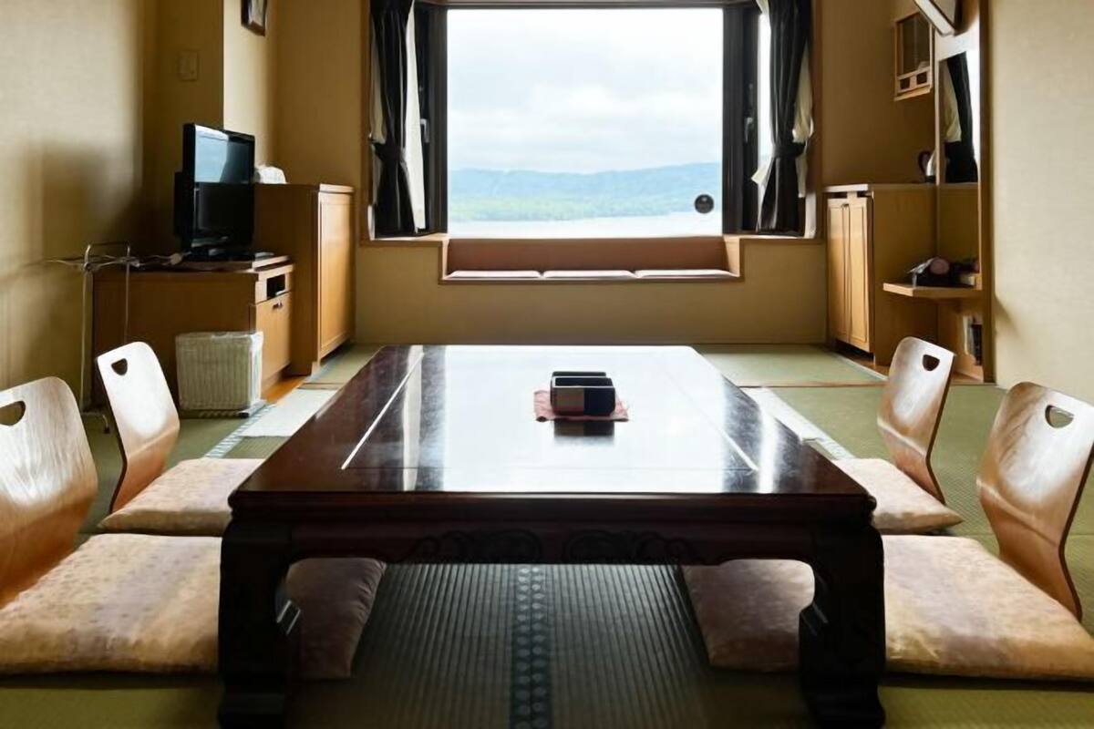 A room with a wooden table, cushioned chairs, a television on a cabinet, and a window with a view of the outdoors.