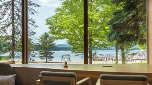 Una habitación con un ventanal grande que da a un lago y una playa, muebles de madera y una mesita con una vela.