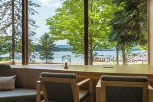 Una habitación con un ventanal grande que da a un lago y una playa, muebles de madera y una mesita con una vela.