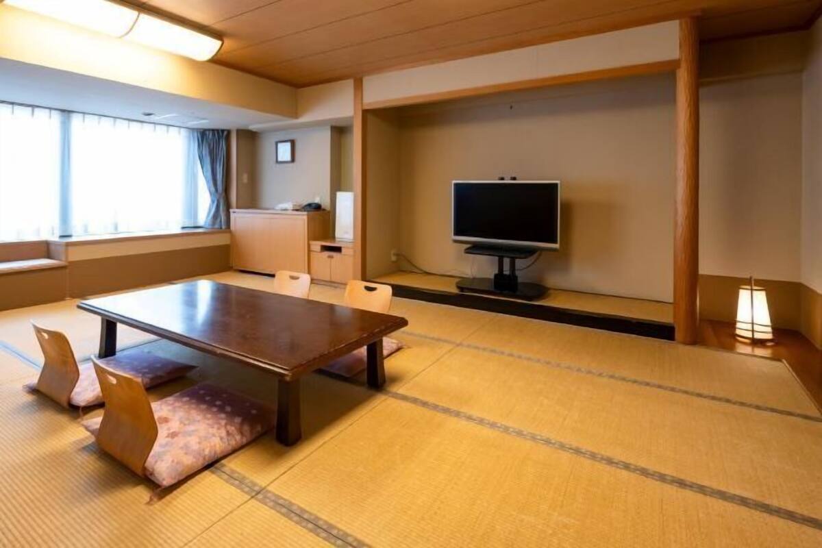 A room with tatami flooring, a low table with cushions, a television on a stand, and a window with curtains.