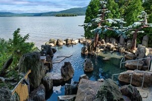 Una fuente termal natural rodeada de rocas y árboles, con vistas a un lago y colinas al fondo.
