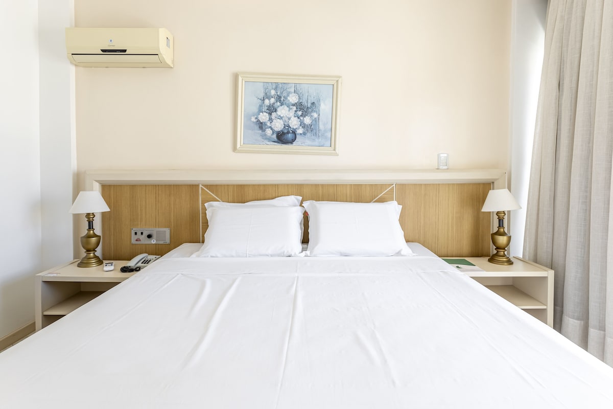 A hotel room with a bed, two bedside tables, a wall-mounted air conditioner, a painting, and a curtain.