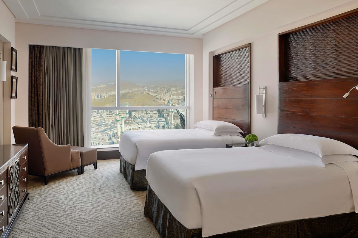 A hotel room with two beds, a desk, a chair, and a large window offering a city view.