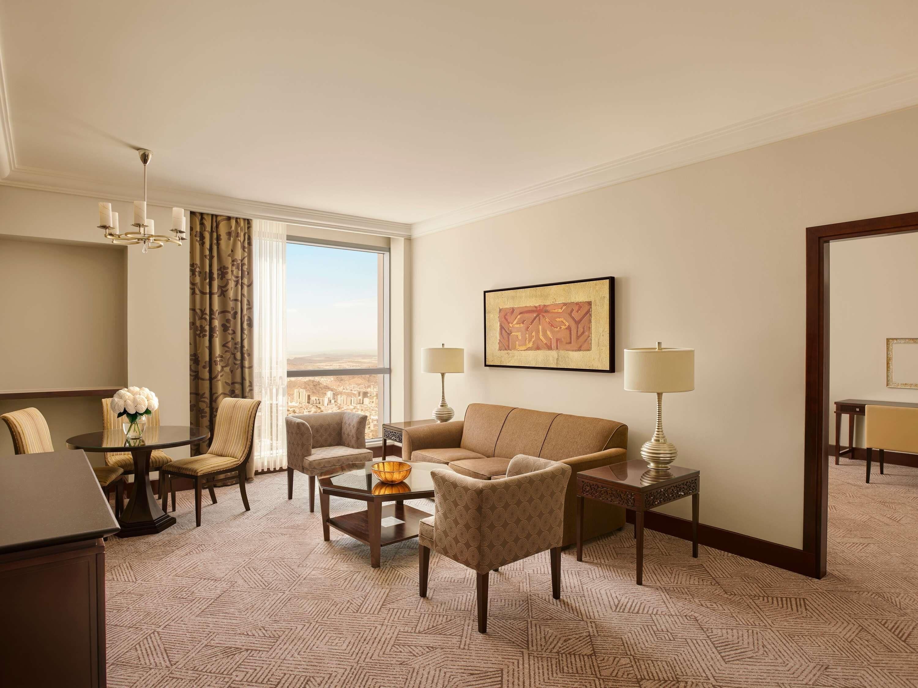 A hotel room with a dining table, two armchairs, a sofa, a coffee table, a lamp, a framed artwork, and a large window with a city view.