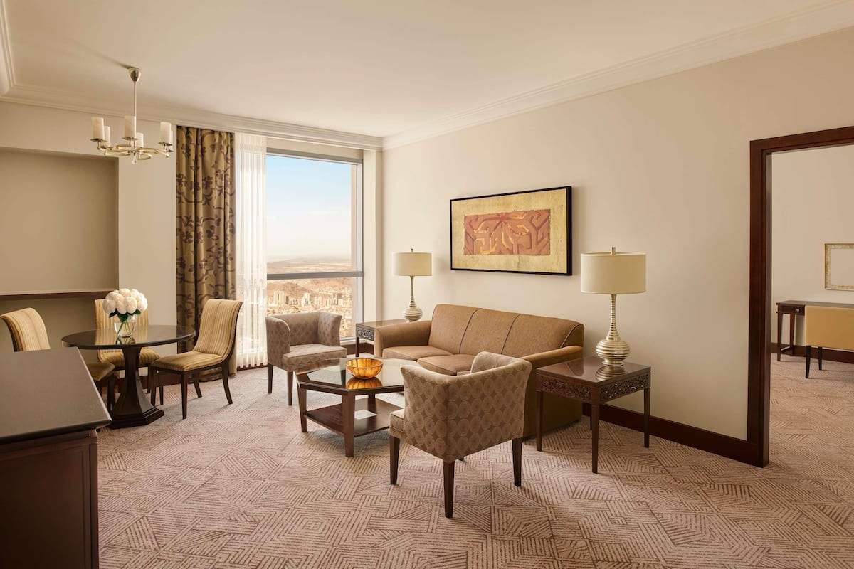 A hotel room with a dining table, two armchairs, a sofa, a coffee table, a lamp, a framed artwork, and a large window with a city view.