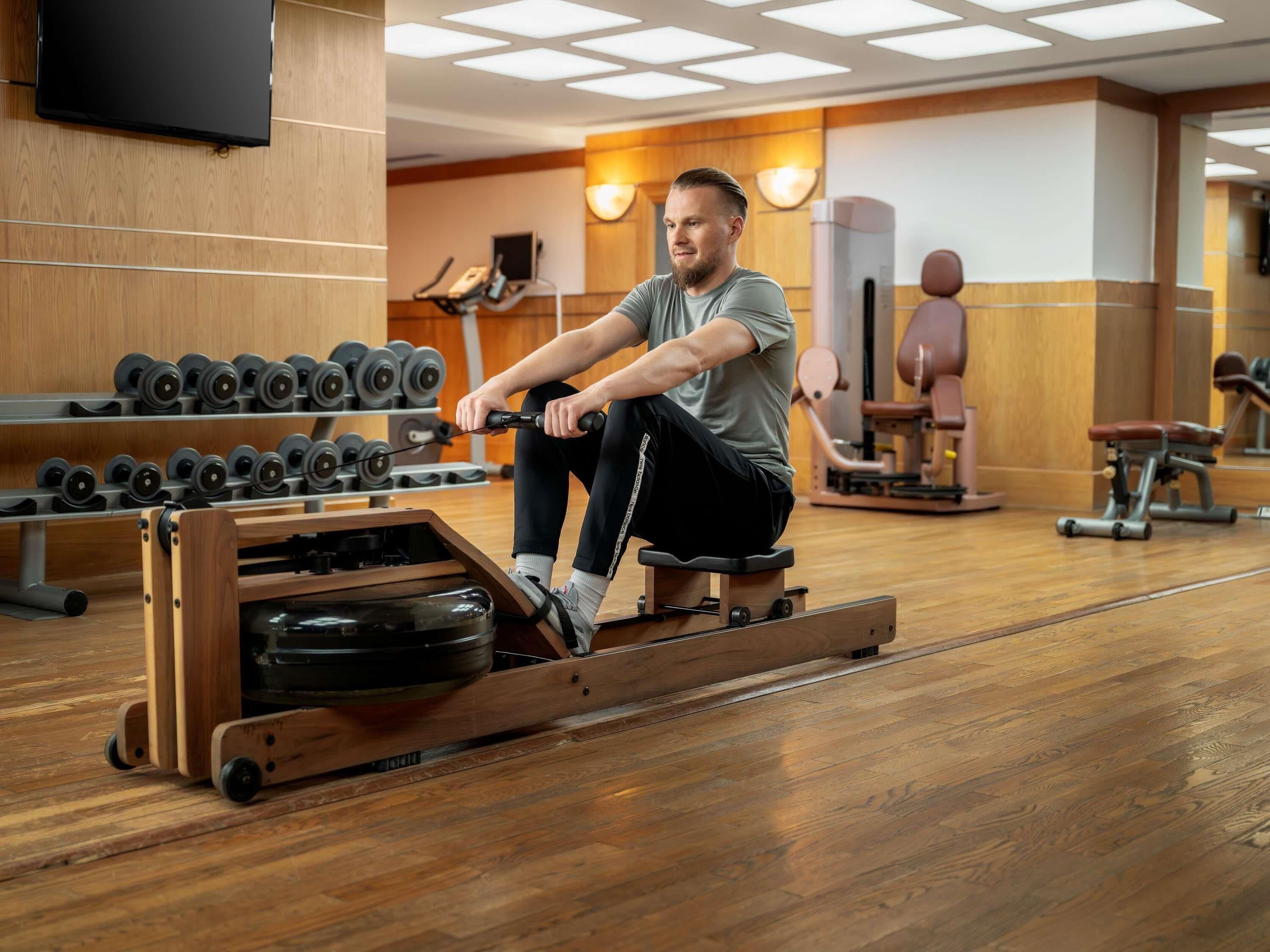 A man using a rowing machine in a gym with wooden flooring, exercise equipment, and a TV mounted on the wall.
