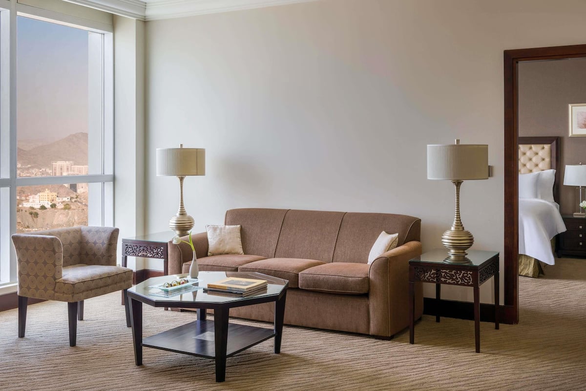 A hotel room with a sofa, armchair, coffee table, two lamps, and a view of a cityscape.