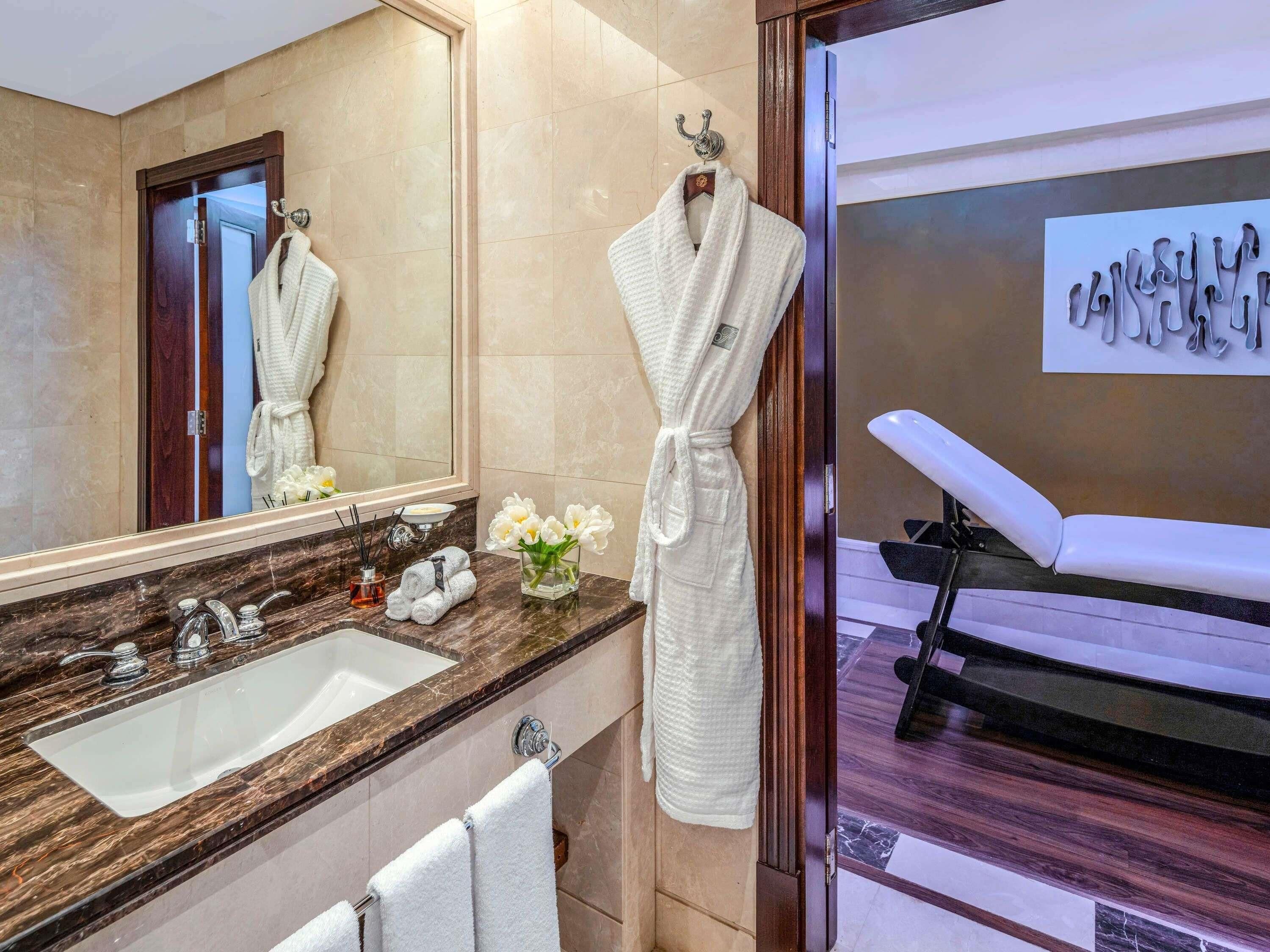Une salle de bain avec un lavabo en marbre, un miroir, un peignoir suspendu à un crochet et un fauteuil de massage.