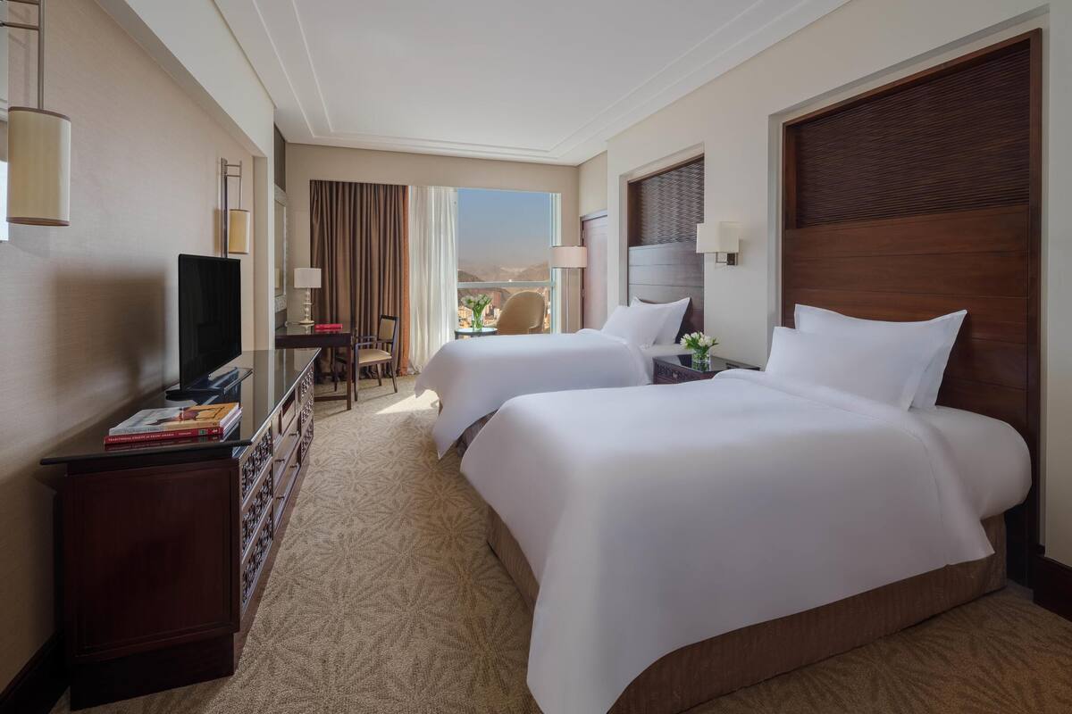 A hotel room with two beds, a desk, a TV, and a view of the city.