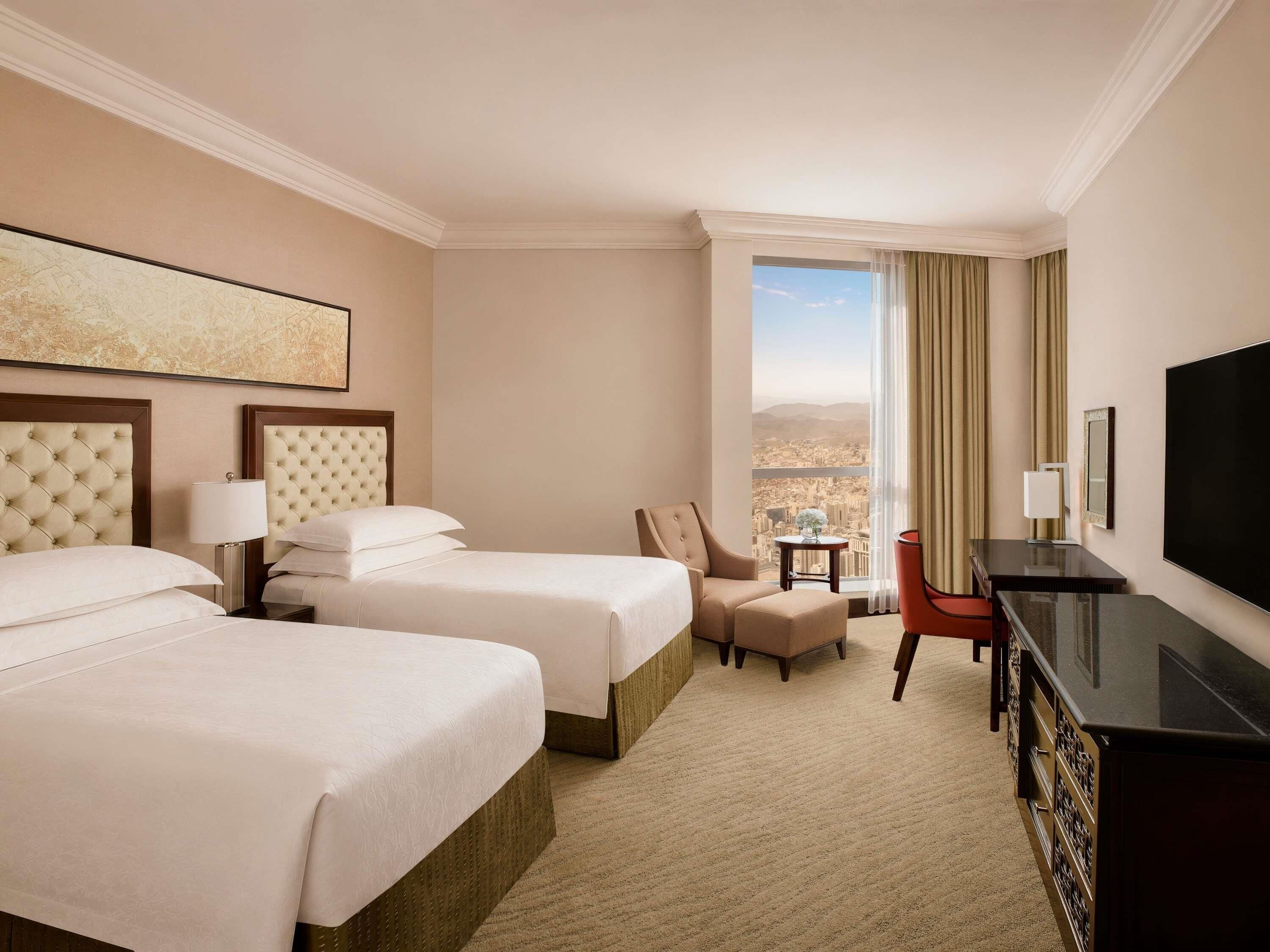 A hotel room with two beds, a desk, a chair, a TV, and a large window with a city view.