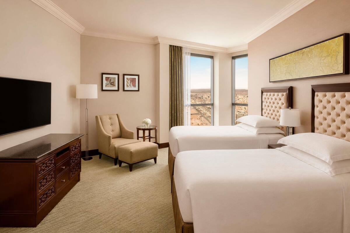 A hotel room with a flat-screen TV, two beds, a chair, a small table, and a window with a city view.
