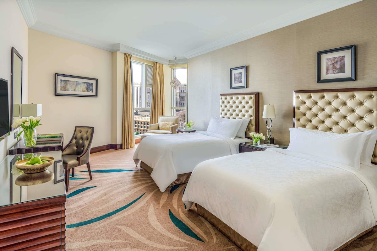 A hotel room with two beds, a desk, a chair, and a view of the city.